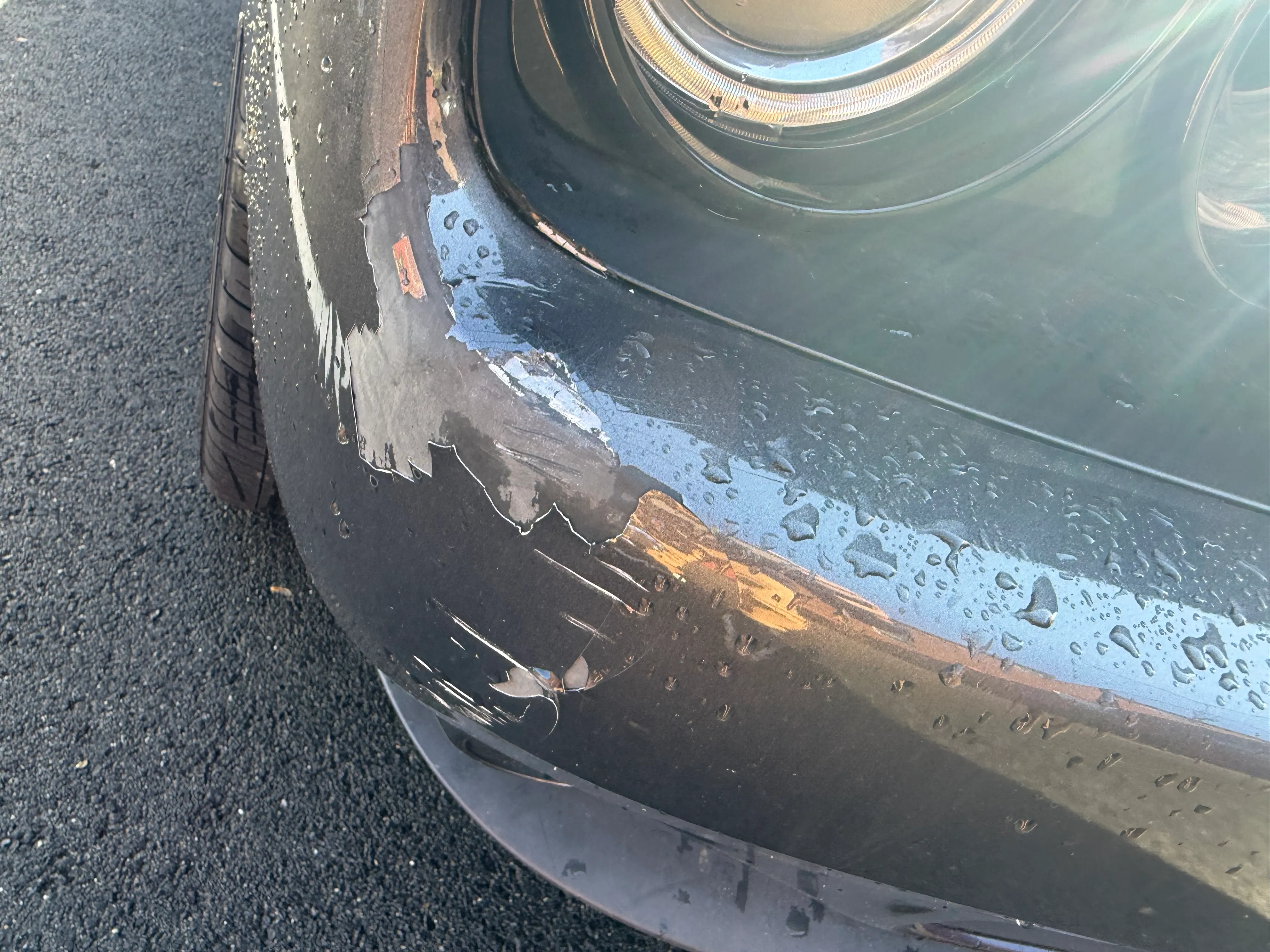 Gray Dodge Challenger front bumper corner with paint and clear coat peeling near the headlight, before repair.