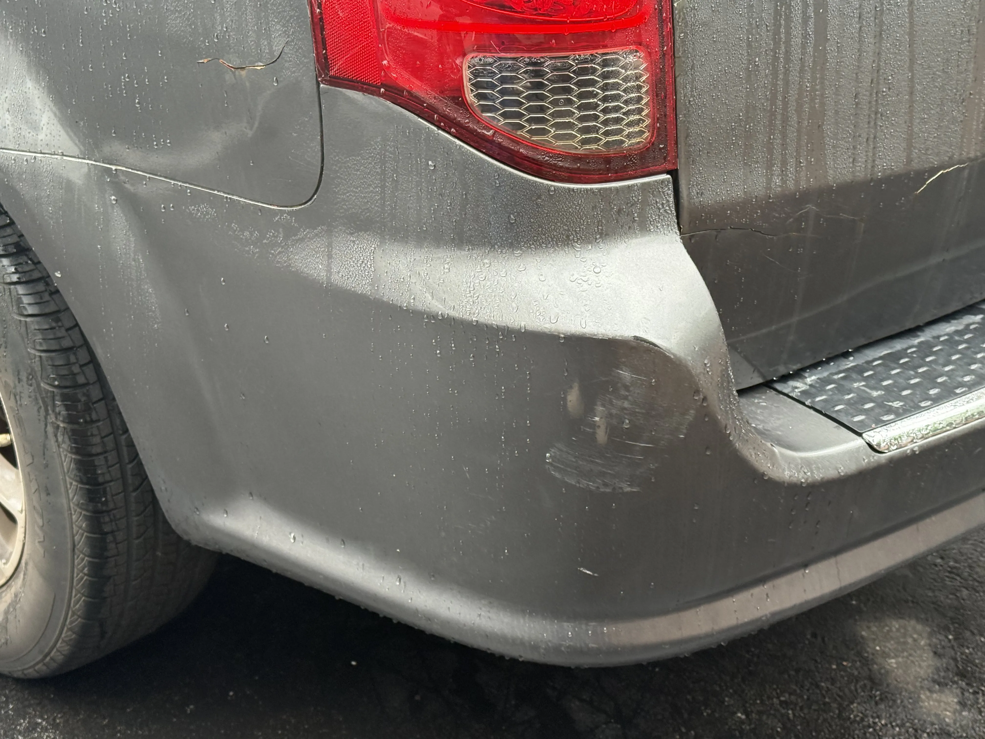 Close-up of gray Dodge Grand Caravan rear bumper with visible dent damage and paint scrape near the taillight, before repair.